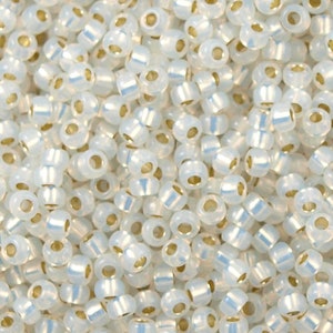 May include: A close-up view of a pile of white seed beads with iridescent shimmer. The beads have a small hole in the center and are approximately 2mm in diameter.