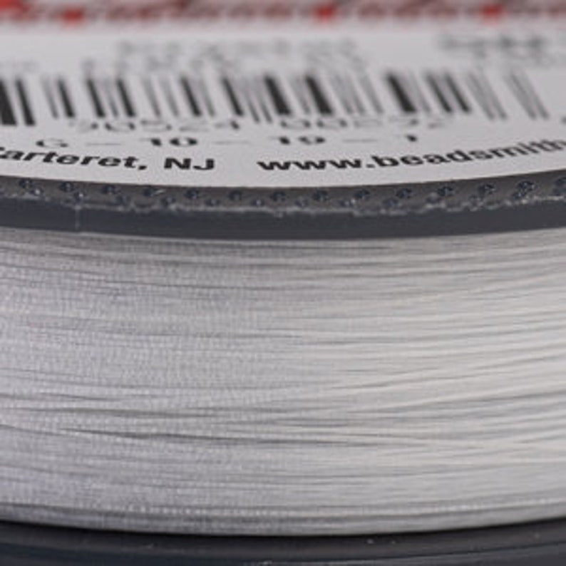 May include: Close-up of a spool of clear, fine beading thread. The thread is tightly wound on a gray spool. The label shows a barcode and text including "Carteret, NJ" and "www.beadsmith.com".