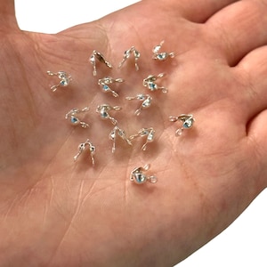 Könnte beinhalten: Nahaufnahme eines Stapels silberfarbener Metall-Crimp-Endkappen für die Schmuckherstellung. Die Kappen haben an einem Ende eine kleine Schlaufe und am anderen Ende einen Crimpbereich.