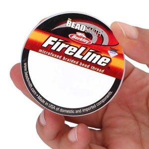 Può includere: Una bobina di filo per perline intrecciato microfuso FireLine. L'etichetta è circolare con un design sfumato rosso, arancione e nero. Il testo "FireLine" è ben visibile, con i loghi Beadsmith e Berkley sopra.