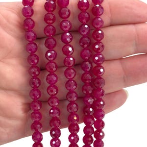 May include: Close-up of several strands of faceted, deep magenta gemstone beads. The round beads have multiple facets, creating a sparkling effect. The beads are strung together, ready for jewelry making. The background is a light colour.