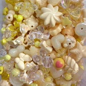 Pode incluir: Uma coleção de várias contas em tons de amarelo, creme e transparente. As contas incluem formas como flores, estrelas, corações, laços e contas redondas. Algumas contas têm um brilho perolado, e outras são translúcidas. A disposição é densa e visualmente atraente.