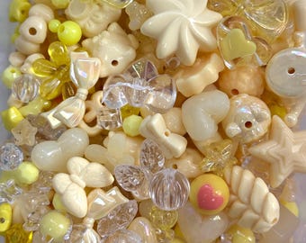 May include: A collection of various beads in shades of yellow, cream, and clear. The beads include shapes like flowers, stars, hearts, bows, and round beads. Some beads have a pearlescent sheen, and others are translucent. The arrangement is dense and visually appealing.
