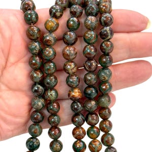 May include: Strands of round beads in shades of brown, green, and gold. The beads have a patterned design and are strung together. The beads are held in a hand against a white background. The beads are approximately 1.3 cm in diameter.