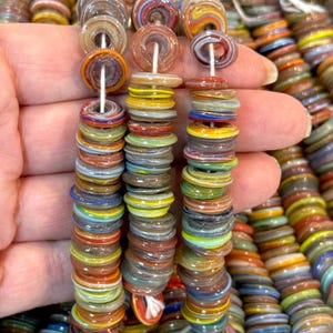 Puede incluir: Primer plano de cuentas de vidrio redondas y planas, multicolores, con tonos de rojo, amarillo, azul y verde. Las cuentas están ensartadas en un hilo blanco y sostenidas en una mano. Las cuentas tienen un diseño rayado y arremolinado.