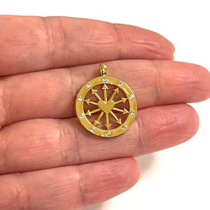May include: Gold pendant with a heart in the center surrounded by arrows pointing outwards. The pendant is set with small clear stones around the edge.
