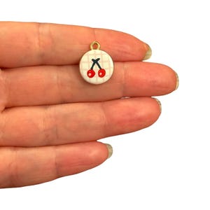 Può includere: Un piccolo ciondolo rotondo in metallo color oro con sfondo bianco e motivo a quadretti rosa chiaro. Al centro, un'illustrazione grafica di due ciliegie rosso brillante con gambi scuri, fissato a un piccolo anello di sospensione dorato.