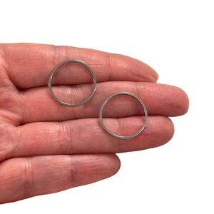 May include: Two silver-colored, thin metal circles. The circles are held in a hand against a white background. The circles are the same size and appear to be jewelry components or findings.