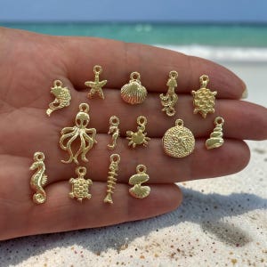 May include: A collection of fourteen small gold-toned metal charms featuring various marine life shapes, including an octopus, seahorses, a turtle, a starfish, a crab, a seashell, a sand dollar, and a fish skeleton, each with a small loop for attachment.