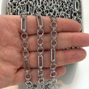 May include: A silver-toned metal chain featuring a repeating pattern of elongated rectangular links interspersed with pairs of small circular links. The chain is constructed from polished, reflective metal segments that create a continuous, uniform design.