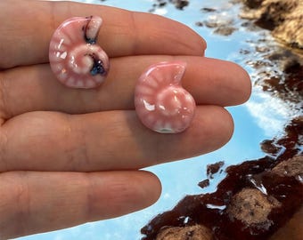 May include: Two small, pink ceramic seashell-shaped objects. The shells have a spiral design and a glossy finish. One shell has a dark blue and black accent. The objects are displayed against a background of water and rocks.