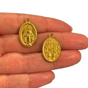 May include: Two gold-coloured oval pendants. One pendant features a detailed depiction of a religious figure. The other pendant displays a cross, the letter M, and stars. Both pendants have a small loop at the top for hanging.