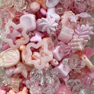 May include: A selection of pink and clear beads in diverse forms. The collection features flower, star, and geometric shapes, alongside translucent and iridescent beads. The beads are in shades of pink, ranging from pale to deeper tones.