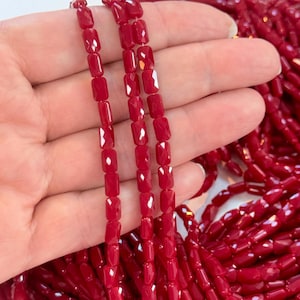 Puede incluir: Una colección de cuentas cilíndricas de color rojo vibrante, facetadas y ensartadas en múltiples hilos. Cada cuenta presenta una superficie pulida y reflectante con facetas geométricas distintivas que captan la luz, creando un aspecto uniforme.
