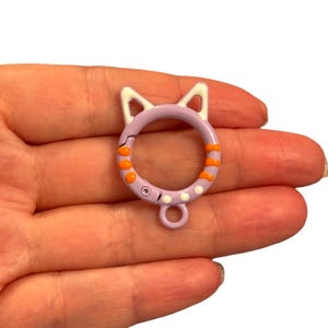 Op de afbeelding: Een lavendelkleurige katvormige sluiting met witte oren en oranje strepen. De sluiting heeft een cirkelvormig ontwerp met een kleine lus aan de onderkant. Witte stippen en een kleine zilveren klinknagel zijn ook zichtbaar.