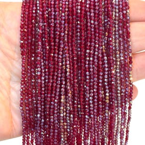 May include: Strands of small, faceted, deep red beads. The beads are strung together, creating a shimmering effect. The beads are held in a hand, showcasing their color and texture. Ideal for jewelry making.