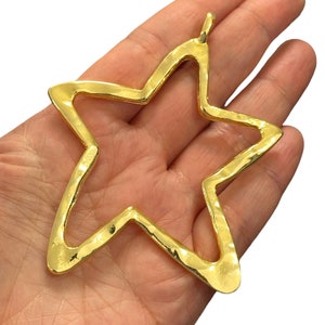 May include: A gold-colored star pendant with a hammered texture. The star has five points and a loop at the top for hanging. The pendant is held in a hand against a white background.