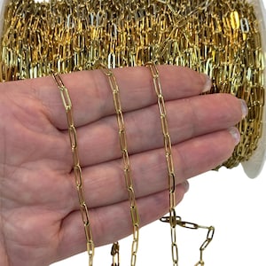 以下が含まれることがあります： 長方形のリンクが付いた金色のチェーンが手で示されています。チェーンはスプールにあり、ジュエリー作りや工芸品に使用されているようです。リンクは均一です。