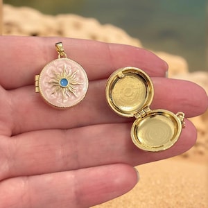 Può includere: Un ciondolo a medaglione color oro con un design a forma di sole in smalto rosa e un centro blu. Il medaglione è aperto, rivelando un interno dorato. Il ciondolo è rotondo e ha un piccolo anello per una catena.