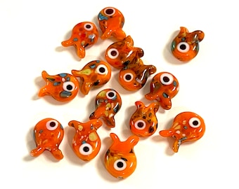 May include: A collection of orange glass fish beads with white and black eyes. The beads have a speckled design with blue, yellow, and brown accents. The fish have a whimsical, cartoonish appearance, and are arranged on a white surface.