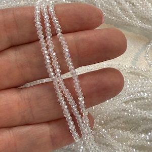 May include: Close-up of several strands of clear, faceted beads. The beads are small and strung together, creating a delicate and shimmering effect. The beads are held in a hand, with more strands visible in the background. The beads are a light, iridescent color.