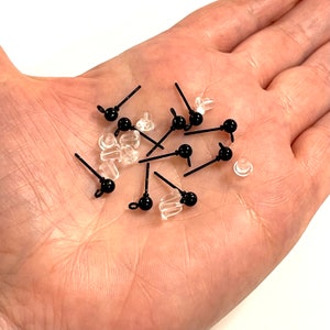 Puede incluir: Primer plano de una mano sosteniendo un montón de pendientes de botón negros con respaldos de plástico transparente. Los pendientes de botón tienen una pequeña bola negra al final del poste.