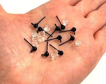 May include: A close-up of a hand holding a pile of black earring studs with clear plastic backs. The earring studs have a small black ball at the end of the post.