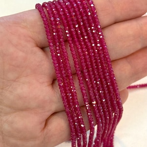 Könnte beinhalten: Mehrere Stränge kleiner, facettierter, tief magentafarbener Perlen. Die Perlen sind gleichmäßig groß und aufgefädelt. Die Perlen haben eine funkelnde, reflektierende Qualität, möglicherweise aus einem Edelstein oder Kristall.