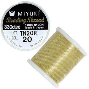 Può includere: Una bobina di filo dorato per perline accanto a un'etichetta rotonda. L'etichetta è in bianco e nero con il testo "MIYUKI Beading Thread" e "330dtex 100% NYLON Made in Japan". La bobina è etichettata "LOT. TN20R COL. 20".