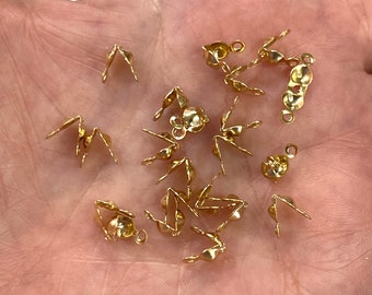 May include: A collection of small, gold-colored jewelry findings, including crimp ends and connector pieces. The findings are scattered across a light-colored surface, showcasing their intricate designs and metallic sheen. These components are used in jewelry making.