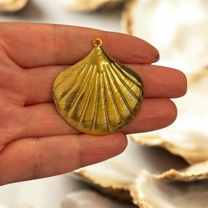 May include: A gold-coloured seashell pendant with a textured surface. The pendant has a loop at the top for hanging and is designed to resemble a scallop shell. The shell has a fan-like pattern.