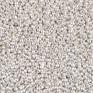 Può includere: Una vista ravvicinata di una grande quantità di perline di rocaille argentate. Le perline sono piccole e rotonde, con un bagliore metallico.