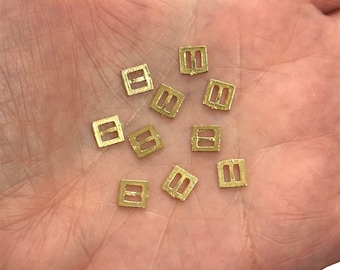 May include: A collection of small, gold-colored metal buckles. The buckles are square-shaped with a rectangular opening in the center.