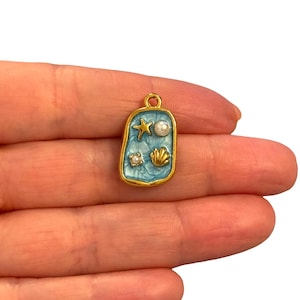 Könnte beinhalten: Ein goldfarbener, rechteckiger Anhänger mit hellblauem Emaille-Hintergrund. Der Anhänger zeigt einen Seestern, eine Perle, eine Muschel und eine kleine Perle. Der Anhänger wird in einer Hand gehalten.