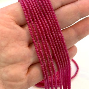Puede incluir: Primer plano de múltiples hebras de pequeñas cuentas redondas de color magenta oscuro. Las cuentas son de tamaño uniforme y están ensartadas. La imagen muestra varias hebras, adecuadas para la fabricación de joyas u otras manualidades.