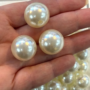 Peut inclure: Gros plan sur trois grosses perles rondes, fausses, de couleur blanc cassé, avec un éclat nacré. Les perles sont lisses et percées d'un petit trou. Les perles sont tenues dans une main.