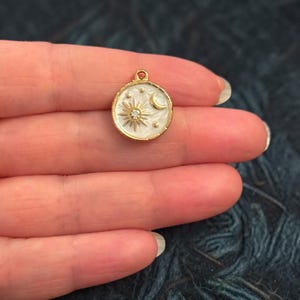 Könnte beinhalten: Goldfarbener Himmelsanhänger mit weißem Emaille-Hintergrund. Der Anhänger zeigt eine Sonne mit einem zentralen Kristall, einen Mond und Sterne. Der Anhänger ist rund und hat oben eine kleine Schlaufe zum Befestigen an Schmuck.