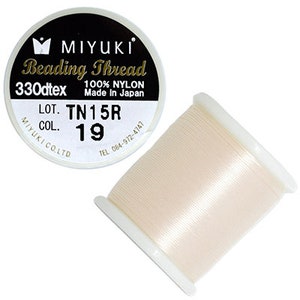 Könnte beinhalten: Eine Spule weißen Miyuki Perlenfadens, 330dtex, 100% Nylon, hergestellt in Japan. Das Etikett lautet "LOT. TN15R COL. 19".
