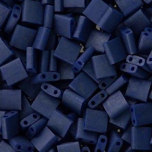 May include: A close-up of a pile of small, dark blue square and rectangular beads. The beads are made of glass or plastic and have a matte finish.