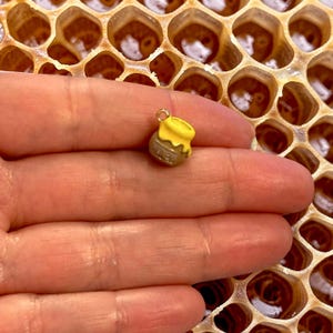 Könnte beinhalten: Ein Miniatur-Honigtopf-Anhänger mit gelbem Deckel und braunem Körper, das Wort "HONEY" ist sichtbar. Der Anhänger wird in einer Hand gehalten, mit einem Bienenwaben-Hintergrund.