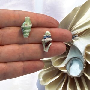 May include: Two miniature ceramic seashell sculptures. One is light green with white and brown accents, the other is white with brown and blue accents. The shells are held in a hand, with a white and cream-coloured flower in the background.