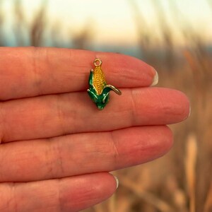 Puede incluir: Un pequeño dije de maíz con un lazo dorado para sujetar a la joyería. El dije presenta una mazorca de maíz amarilla con hojas verdes. El dije mide aproximadamente 2 cm de largo.