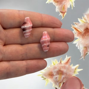 以下が含まれることがあります： 白と茶色のアクセントが付いた、2つの小さなピンク色のセラミック貝殻ビーズ。ビーズは手で持たれており、ピンクと黄色の花の要素がぼやけた背景になっています。