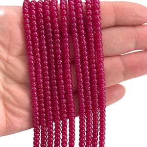 May include: Strands of small, round, deep red beads. The beads are strung together, creating multiple vertical lines. The beads have a glossy finish and are held in a hand against a white background. These beads are suitable for jewelry making.