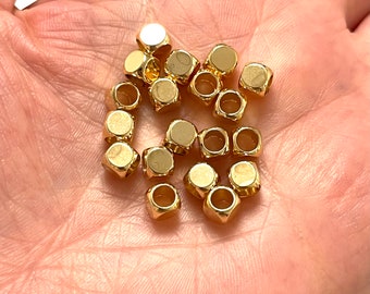 May include: A close-up of a pile of gold-colored metal beads. The beads are round with a flat top and bottom, and have a small hole in the center. The beads are arranged in a random pattern.