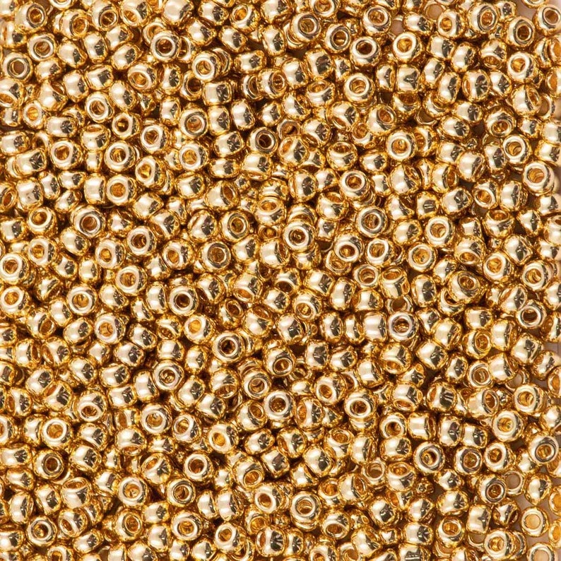 May include: A close-up image of a large quantity of gold-colored seed beads. The beads are round and have a shiny finish.