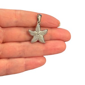 May include: A silver-toned starfish pendant featuring a textured surface covered in small, raised circular dots. The pendant has a smooth, polished bail at the top for attachment to a chain, showcasing a five-pointed star shape with a metallic finish.