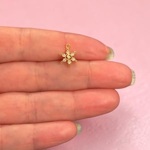 Può includere: Un piccolo ciondolo dorato a forma di stella o fiocco di neve. Il ciondolo è adornato con piccole gemme trasparenti. Il ciondolo è tenuto nella mano di una persona su uno sfondo rosa.