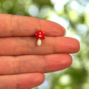 Puede incluir: Un pequeño colgante de hongo rojo con lunares blancos y un tallo blanco. El colgante tiene un lazo dorado en la parte superior para sujetarlo a la joyería. El hongo está en la mano de una persona.