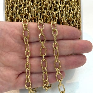 Könnte beinhalten: Eine goldfarbene Metallkette mit ovalen Gliedern, die eine texturierte, gedrehte Oberflächenstruktur aufweisen. Die Kette wird als durchgehende Spule präsentiert, wobei drei Stränge vertikal herabhängen und das sich wiederholende Gliedermuster zeigen.